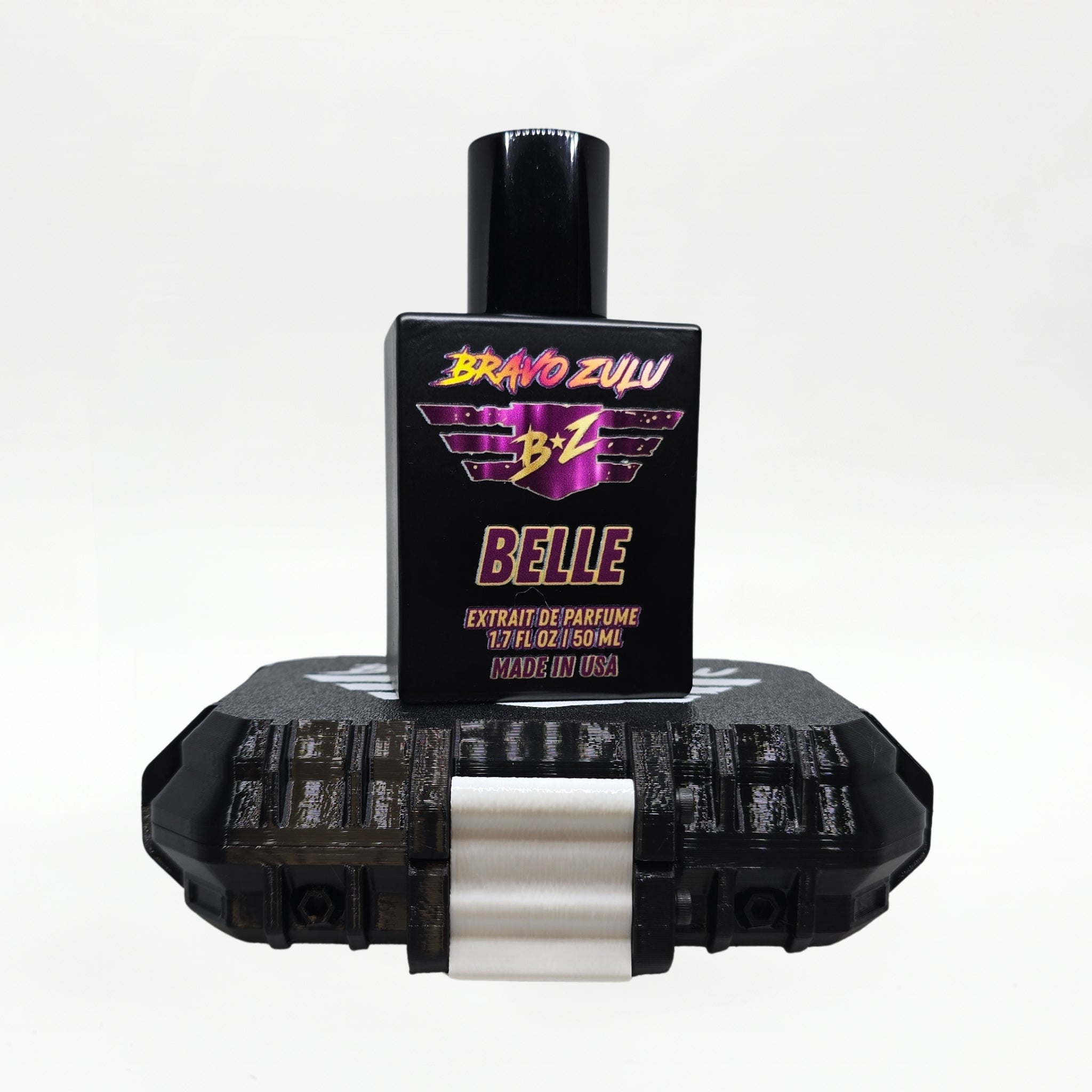 Bravo Zulu BELLE Extrait de Parfum bottle by Flagship Soap Company balanced on top of its closed black rugged tactical case, the matte black glass bottle displaying the maroon-and-gold BRAVO ZULU B★Z winged logo and BELLE name, on a seamless white studio background.