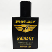 Front-facing studio product shot of Bravo Zulu RADIANT Extrait de Parfum by Flagship Soap Company. The heavy matte black glass bottle with matching black cap is centered on a clean white background, displaying the gold-and-black BRAVO ZULU B★Z winged logo, RADIANT name, Extrait de Parfume 1.7 FL OZ / 50 ML, and Made in USA text.