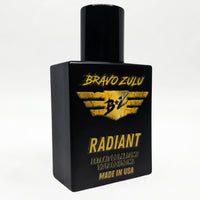 Angled studio shot of the Bravo Zulu RADIANT Extrait de Parfum bottle by Flagship Soap Company, showing the matte black glass bottle and black cap with gold-and-black BRAVO ZULU B★Z winged logo and RADIANT branding, set against a seamless white studio background.
