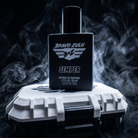 A dynamic, tactical product photograph for SEMPER Extrait de Parfum by Bravo Zulu. A heavy, matte black glass bottle, featuring a distinct white B+Z winged shield logo and bold 'SEMPER' branding, stands centrally on its custom white and black rugged tactical protective case. The complex scene is enveloped in dramatic, cool-toned smoke and mist against a dark, low-key background, visualizing the cold ocean marine and rugged earthy woods scent profile. Key keywords integrated into the visual description inclu