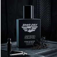 A tactical product image of SEMPER Extrait de Parfum by Bravo Zulu. The heavy matte black glass bottle is positioned on a rugged, weathered armor plate alongside dark metal shell casings and a fresh, cold juniper branch with blue berries, showcasing the scent's rugged earthy woods foundation. The background features a dark carbon fiber texture, capturing an elite, heavy-duty aesthetic engineered for a commanding, unyielding presence.