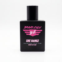 A front-facing, professional product studio shot of the Bravo Zulu Flagship Soap Company — SHE BANGZ Extrait de Parfum (Women's Perfume) bottle, as seen in image_21.png. The fragrance is housed in a heavy, tactical matte black glass bottle with a matching black cylindrical cap, set against a clean, brilliant white seamless background. The front of the bottle features the distinct pink and white branding:

BRAVO ZULU (Top, pink text).

B Z (Centered, pink stylized logo with a pink star).

SHE BANGZ (Centered