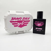 A professional studio product shot featuring the premium hardware of SHE BANGZ Extrait de Parfum from the Bravo Zulu Lineup by Flagship Soap Company. The heavy, tactical matte black glass bottle stands alongside its closed, custom-engineered rugged protective tactical case, both cleanly isolated on a seamless, brilliant white background.

The front of the sleek black bottle displays vibrant pink and white branding text for "BRAVO ZULU" and "SHE BANGZ," noting its high-impact 30% oil concentration and Americ