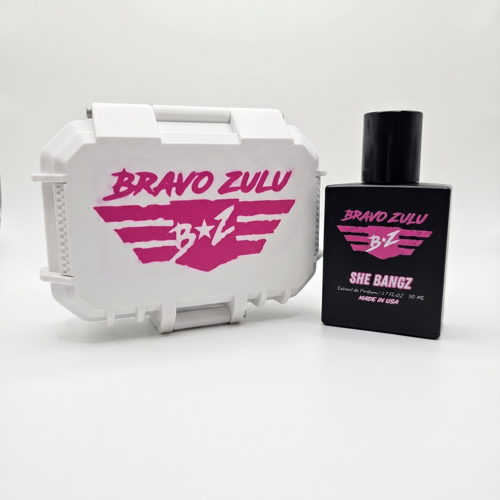 A professional studio product shot featuring the premium hardware of SHE BANGZ Extrait de Parfum from the Bravo Zulu Lineup by Flagship Soap Company. The heavy, tactical matte black glass bottle stands alongside its closed, custom-engineered rugged protective tactical case, both cleanly isolated on a seamless, brilliant white background.

The front of the sleek black bottle displays vibrant pink and white branding text for "BRAVO ZULU" and "SHE BANGZ," noting its high-impact 30% oil concentration and Americ