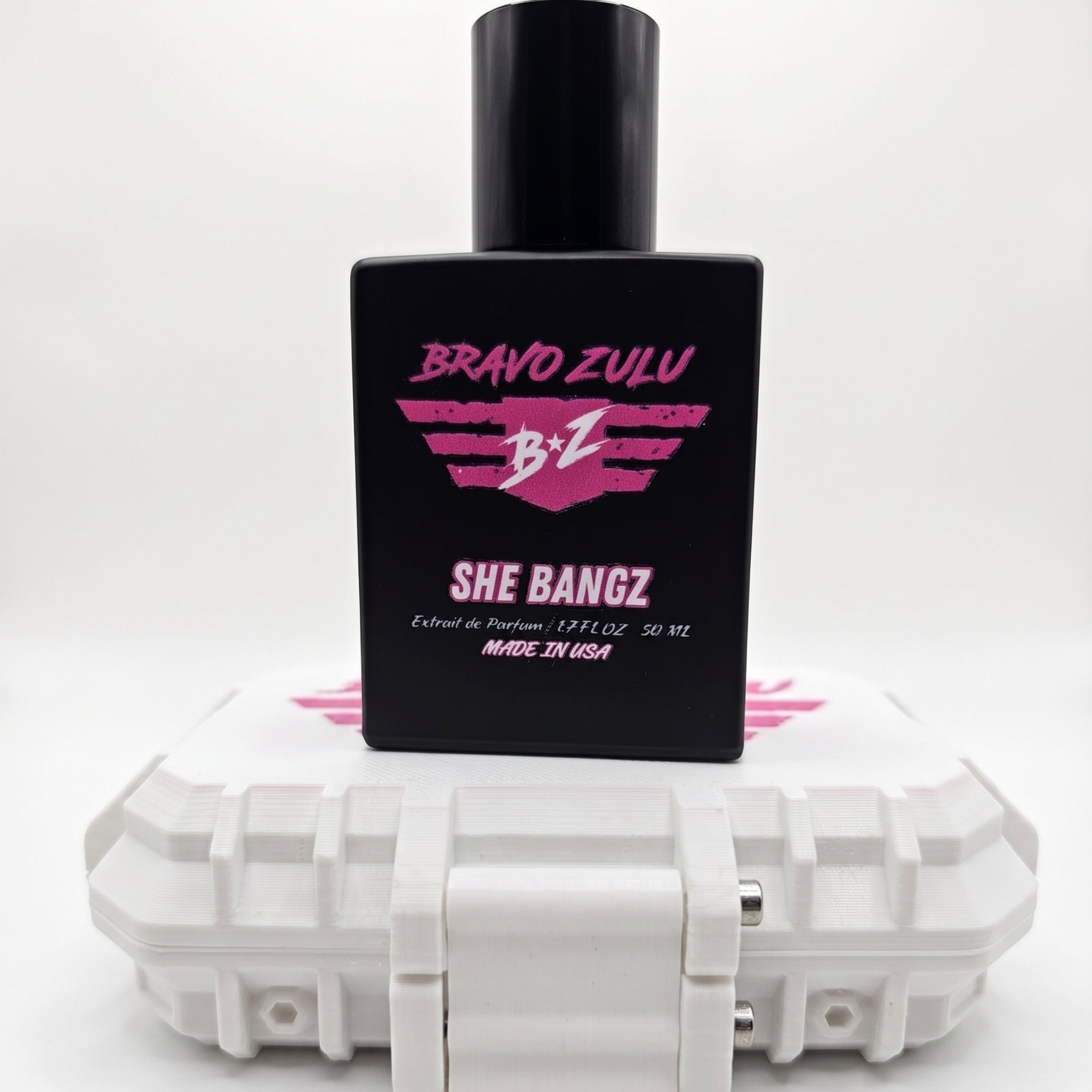 Shopify product image of SHE BANGZ Extrait de Parfum from the Bravo Zulu Lineup by Flagship Soap Company. The heavy, tactical matte black glass bottle is prominently balanced and displayed sitting on top of its closed, custom-engineered rugged protective tactical case. Isolated against a clean, seamless, brilliant white studio background, the sleek black bottle highlights the vibrant hot pink and white branding for "BRAVO ZULU" and "SHE BANGZ." The label reflects its premium, high-impact 30% oil concentrati