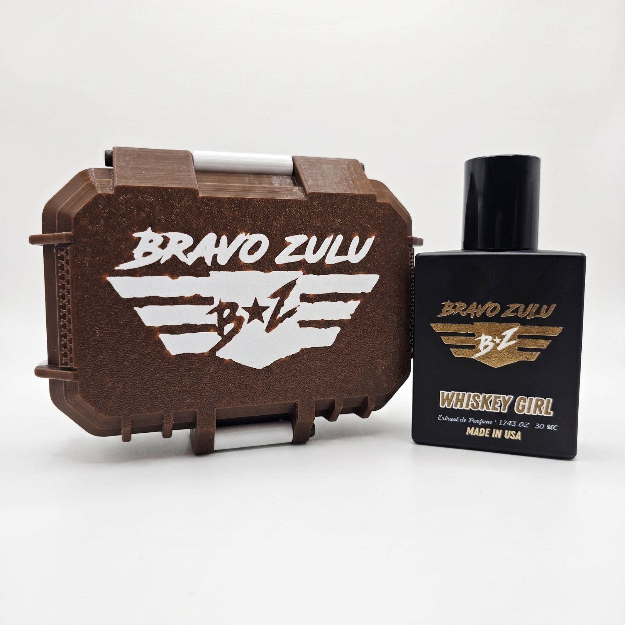 Front view of Whiskey Girl Extrait de Parfum by Flagship Soap Company, displayed next to its custom case on a clean white background. On the right, the tactical matte black glass bottle features a gold-embossed Bravo Zulu wings logo and "WHISKEY GIRL" label. On the left, the closed, rugged tactical case is finished in Bourbon Brown with a bold white-embossed Bravo Zulu flight wing emblem across the lid.