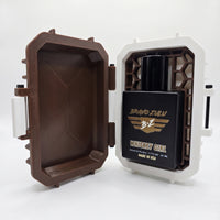An open view of the Whiskey Girl Extrait de Parfum by Flagship Soap Company securely docked in its custom case. The heavy, tactical matte black glass bottle sits inside the white contrast frame, showcasing a textured brown honeycomb insert that holds the bottle upright. The left side displays the open, rugged Bourbon Brown lid, shot against a seamless white background.
