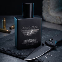 Bravo-Zulu-Flagship-Soap-Company-Extrait-De-Parum-Fragrance-Cologne-Bottle-Knife.jpg depicts the compact 40ml matte black glass bottle of Bravo Zulu Flagship Soap Company's BLACKOUT Extrait de Parfum. The heavy, tactical masterpiece sits on a textured slate surface, its monochromatic Stealth Black design featuring silver embossed 'BRAVO ZULU' and 'BLACKOUT' logos, and 'MADE IN USA' text, all engineered for the men who defend the perimeter. Next to it, a black tactical folding knife with a textured grip and 