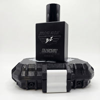 A clean studio product shot on a white background showcasing the BLACKOUT Extrait de Parfum by Flagship Soap Company. The compact footprint matte black glass bottle is positioned front and center, standing balanced on top of its rugged, custom-engineered tactical case. The bottle features the embossed 'BRAVO ZULU' text and winged 'B+Z' logo, with white text reading 'BLACKOUT', 'COLOGNE | 1.7 FL OZ | 50 ML', and 'MADE IN USA'. The rugged, heavy-duty protective case beneath it features a monochromatic black b