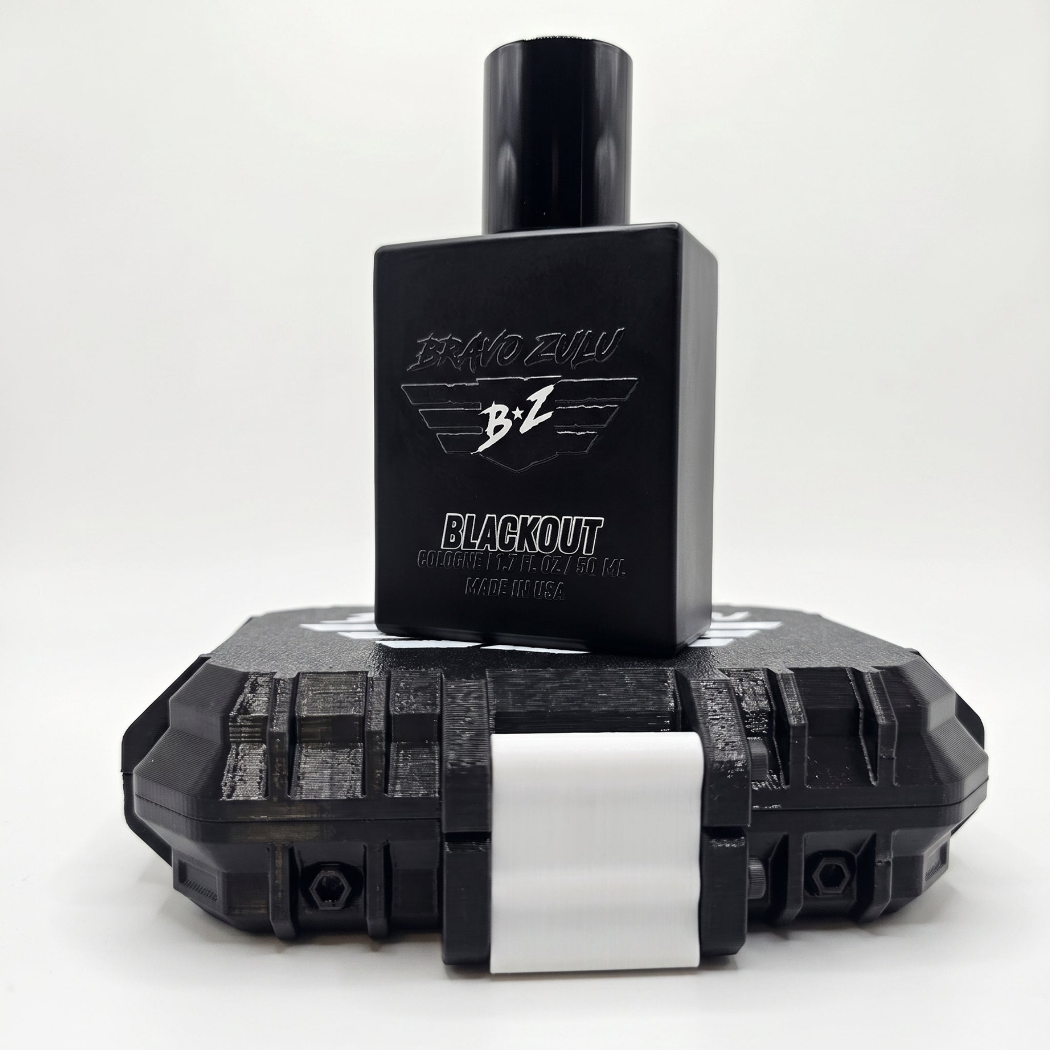 A clean studio product shot on a white background showcasing the BLACKOUT Extrait de Parfum by Flagship Soap Company. The compact footprint matte black glass bottle is positioned front and center, standing balanced on top of its rugged, custom-engineered tactical case. The bottle features the embossed 'BRAVO ZULU' text and winged 'B+Z' logo, with white text reading 'BLACKOUT', 'COLOGNE | 1.7 FL OZ | 50 ML', and 'MADE IN USA'. The rugged, heavy-duty protective case beneath it features a monochromatic black b