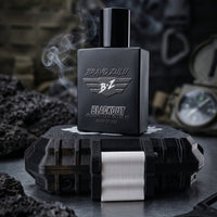 A gritty, atmospheric product shot featuring the BRAVO ZULU BLACKOUT cologne bottle standing upright on its closed tactical case against a dark, rugged background. Wisps of white smoke curl upward past the heavy matte black glass bottle, visually capturing the fragrance's intoxicating smoke screen profile of pitch-black musk, rugged woods, and heavy spices. The bottle prominently displays the embossed 'BRAVO ZULU' text and winged 'B+Z' logo, alongside silver text for 'BLACKOUT COLOGNE', '1.7 FL OZ / 50 ML',