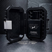 An e-commerce product shot displaying the premium unboxing experience of the BLACKOUT Extrait de Parfum by Flagship Soap Company. The custom-engineered tactical case is wide open on a dark metallic table, revealing its rugged interior with a specialized honeycomb matrix structure on the right side that securely locks the heavy, tactical matte black glass bottle in place. The bottle features clean silver branding including the 'BRAVO ZULU' text, the signature 'B+Z' winged logo, and text below reading 'BLACKO