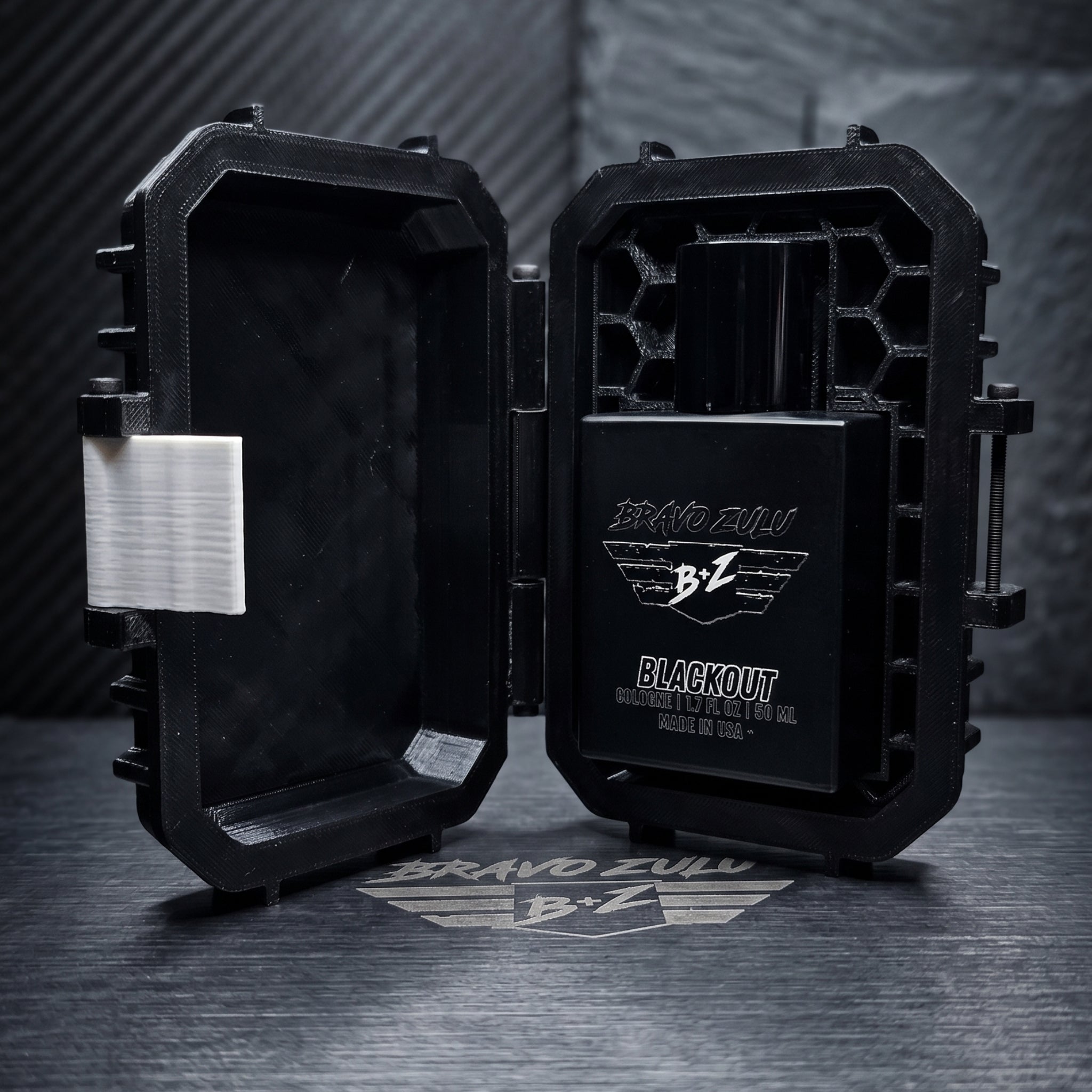 An e-commerce product shot displaying the premium unboxing experience of the BLACKOUT Extrait de Parfum by Flagship Soap Company. The custom-engineered tactical case is wide open on a dark metallic table, revealing its rugged interior with a specialized honeycomb matrix structure on the right side that securely locks the heavy, tactical matte black glass bottle in place. The bottle features clean silver branding including the 'BRAVO ZULU' text, the signature 'B+Z' winged logo, and text below reading 'BLACKO