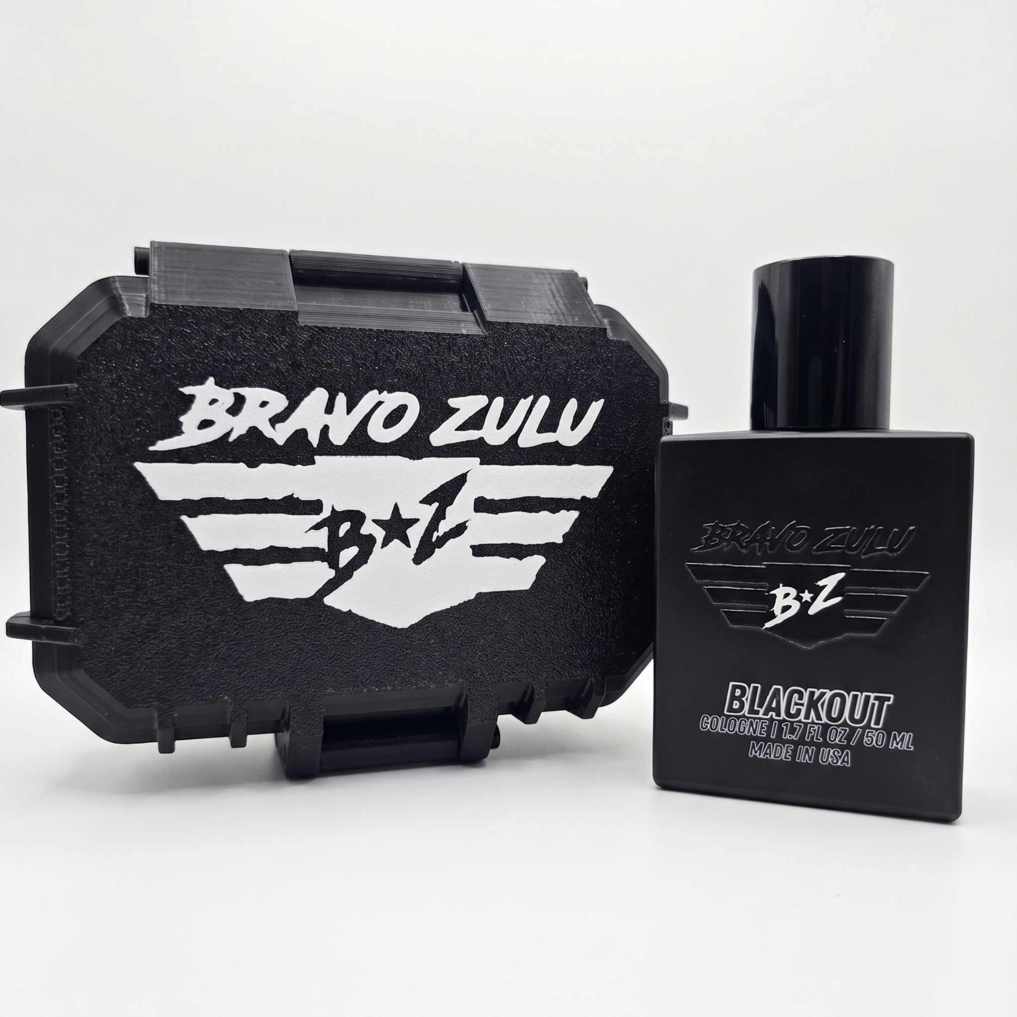A clean e-commerce studio product shot on a crisp white background showing a side-by-side view of the premium hardware included with BLACKOUT Extrait de Parfum by Flagship Soap Company. On the left sits the closed, rugged custom-engineered stealth black tactical case featuring a prominent white embossed 'BRAVO ZULU' title and signature 'B+Z' winged logo design. Standing to the right is the heavy, tactical matte black glass bottle featuring an all-black cap. The front of the bottle displays the matching glos