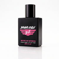 An angled view of the heavy tactical matte black glass bottle of Bravo Zulu MERICAN WOMAN Extrait de Parfum, standing on a clean white reflective surface. The bottle features a striking elite pink and white wings logo and pink typography displaying the product name. This 30% concentration premium feminine fragrance is American-built and engineered with sweet confectionary fruits, exotic midnight florals, and rich vanilla amber.