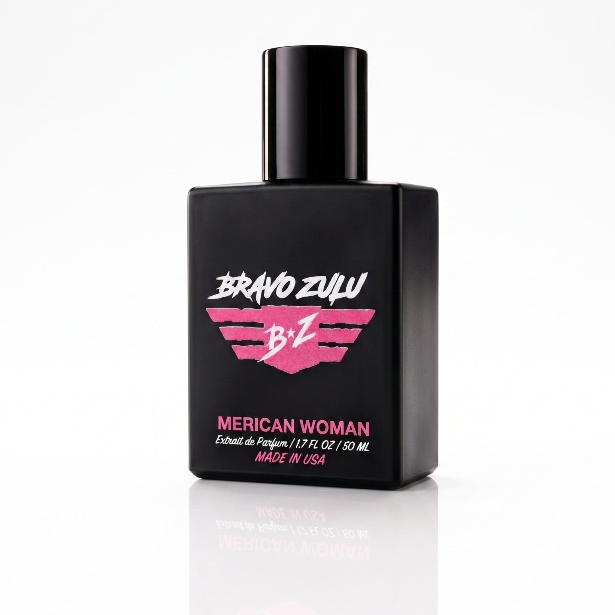 An angled view of the heavy tactical matte black glass bottle of Bravo Zulu MERICAN WOMAN Extrait de Parfum, standing on a clean white reflective surface. The bottle features a striking elite pink and white wings logo and pink typography displaying the product name. This 30% concentration premium feminine fragrance is American-built and engineered with sweet confectionary fruits, exotic midnight florals, and rich vanilla amber.