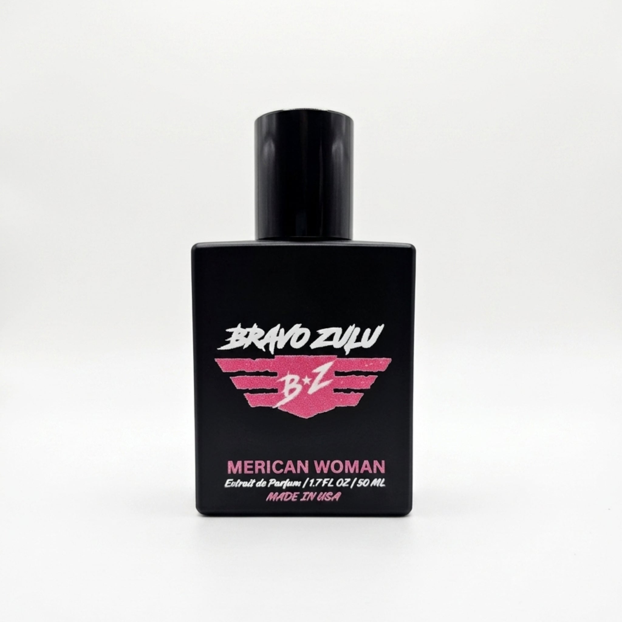 A straight, front-facing product shot of the Bravo Zulu MERICAN WOMAN Extrait de Parfum perfume bottle centered on a completely seamless, clean white background. The heavy, tactical matte black glass bottle is labeled with a bold pink and white winged B★Z logo and pink typography displaying the name and technical specifications. Formulated at a high-octane 30% fragrance oil concentration and proudly made in the USA, this premium blend features scent notes of sweet confectionary fruits, exotic midnight flora