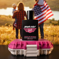 A front-focused product shot of the Bravo Zulu MERICAN WOMAN Extrait de Parfum perfume bottle balanced on top of its locked pink and white tactical case. In the softly blurred, sunlit outdoor background, a family stands in a golden field near a lake, with a woman wearing flannel carrying a child and a man holding an American flag. The matte black glass bottle cleanly displays the bold pink and white winged B★Z logo. Formulated at a high-octane 30% fragrance oil concentration and made in the USA, this premiu