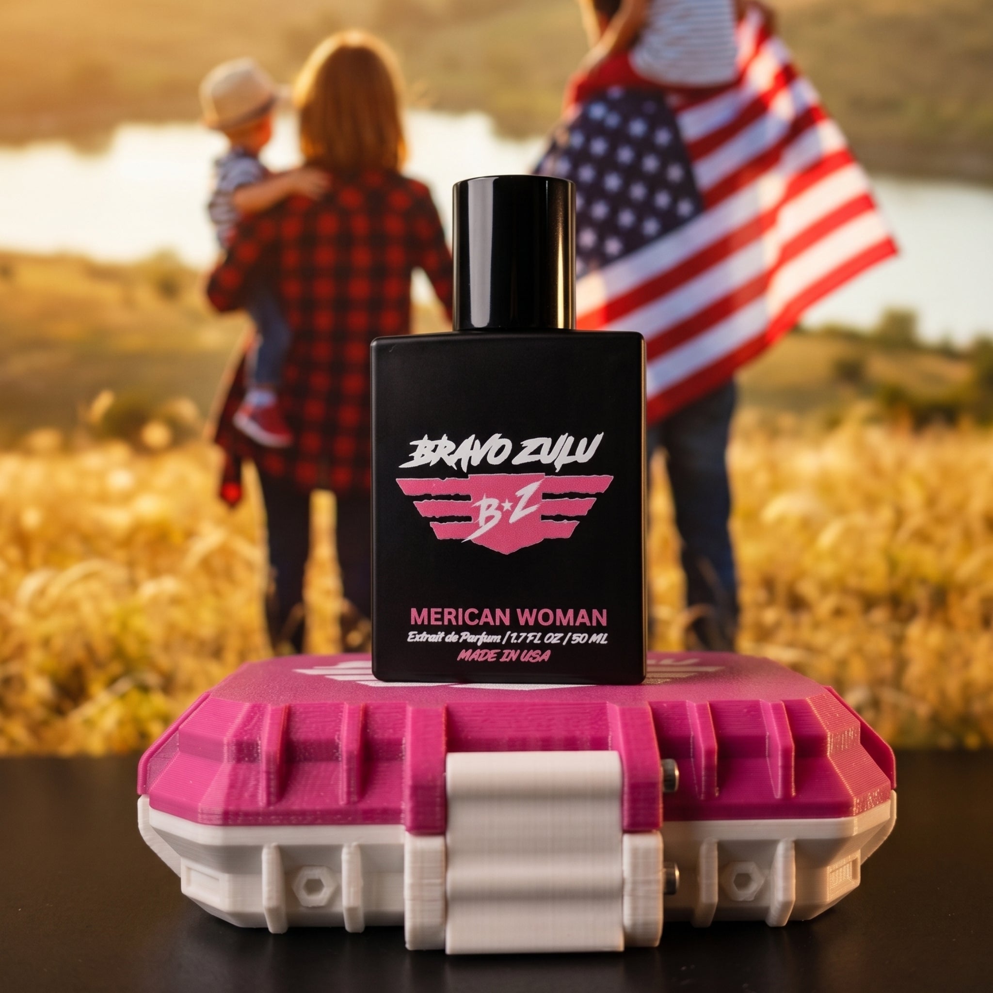 A front-focused product shot of the Bravo Zulu MERICAN WOMAN Extrait de Parfum perfume bottle balanced on top of its locked pink and white tactical case. In the softly blurred, sunlit outdoor background, a family stands in a golden field near a lake, with a woman wearing flannel carrying a child and a man holding an American flag. The matte black glass bottle cleanly displays the bold pink and white winged B★Z logo. Formulated at a high-octane 30% fragrance oil concentration and made in the USA, this premiu
