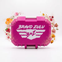 A centered, front-facing product shot of the closed Bravo Zulu MERICAN WOMAN custom tactical protective case on a crisp white studio background. The rugged, custom-engineered case is finished in a striking Elite Pink with clean white contrast latches and features the white winged B★Z logo. Floating dynamically around the hardware are physical representations of its scent architecture: juicy Florida tangerines, tart red berries, a pink bubblegum sphere, exotic midnight orchids, and warm golden amber crystals