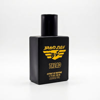 Three-quarter angled view of the TREAD Extrait de Parfum cologne bottle from Flagship Soap Company, isolated on a seamless white background. The heavy, tactical matte black glass bottle is turned slightly to show its dimensional, clean-lined footprint and deep black cap. The front displays the aggressive strike yellow "Bravo Zulu" winged logo, the product name "TREAD", and printed gold text at the base reading "EXTRAIT DE PARFUME | 1.7 FL OZ | 50 ML | MADE IN USA".