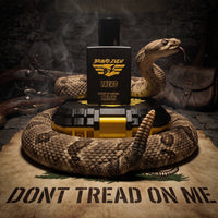 High-impact, tactical product shot for TREAD Extrait de Parfum by Flagship Soap Company. The heavy, matte black glass cologne bottle stands upright on top of its closed custom tactical case, which features an aggressive matte black top and high-visibility strike yellow base. A highly detailed, realistic rattlesnake coils completely around the base of the setup, extending its head with its tongue out to the right of the bottle, while its rattle is raised prominently in the center foreground. The bottle featu