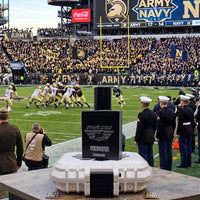 Alt Text: A high-impact product display featuring the Bravo Zulu VENDETTA cologne bottle and its custom rugged protective case centered in the foreground, set against the backdrop of the iconic Army-Navy football game stadium rivalry.

The heavy, tactical matte black glass bottle stands upright on top of its white protective case, which is positioned on a wooden table. The silver-embossed label on the front of the hardware displays the "BRAVO ZULU B★Z" winged shield insignia and the bold title text "VENDETT