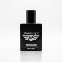 A high-resolution product photograph on a white background with a sharp reflection, featuring the Bravo Zulu VENDETTA Extrait de Parfum (cologne) bottle. The bottle is a matte black glass container with a glossy black cylindrical cap, designed with American grit and resilience for the man who handles business with calculated precision. The silver label on the front features the text "BRAVO ZULU" at the top, a stylized winged shield emblem with "B Z" on it, and "VENDETTA" in larger text below the emblem. At 