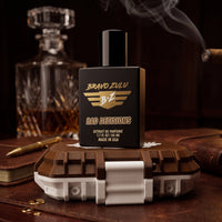 Product image of Bravo Zulu BAD DECISIONS 40ml men's cologne by Flagship Soap Company. The tactical matte black glass bottle sits on its rugged brown and white protective case in a speakeasy lounge setting, capturing the warm, boozy essence of aged barrel-proof bourbon, charred white oak, rich vanilla bean, and deep masculine musk with a 30% fragrance oil concentration.