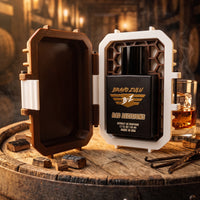 An open brown and white custom protective case holding a matte black bottle of Bravo Zulu BAD DECISIONS Extrait de Parfum. Poured by hand in the USA with a commercial strength 30% oil concentration, the whiskey-themed men's fragrance is presented on a rustic oak barrel head next to a glass of bourbon and vanilla pods inside an atmospheric distillery aging cellar.