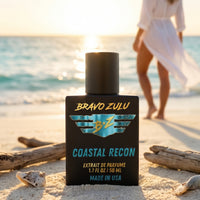 Lifestyle product shot of Bravo Zulu COASTAL RECON Extrait de Parfum by Flagship Soap Company. The matte black glass bottle with turquoise-and-gold BRAVO ZULU B★Z winged logo and COASTAL RECON branding sits in sharp focus on a sunlit beach among driftwood and seashells, with a woman in a flowing white coverup walking along the shoreline at golden-hour sunset in the softly blurred background, evoking the fragrance's bright coconut-pineapple-white-floral coastal character.