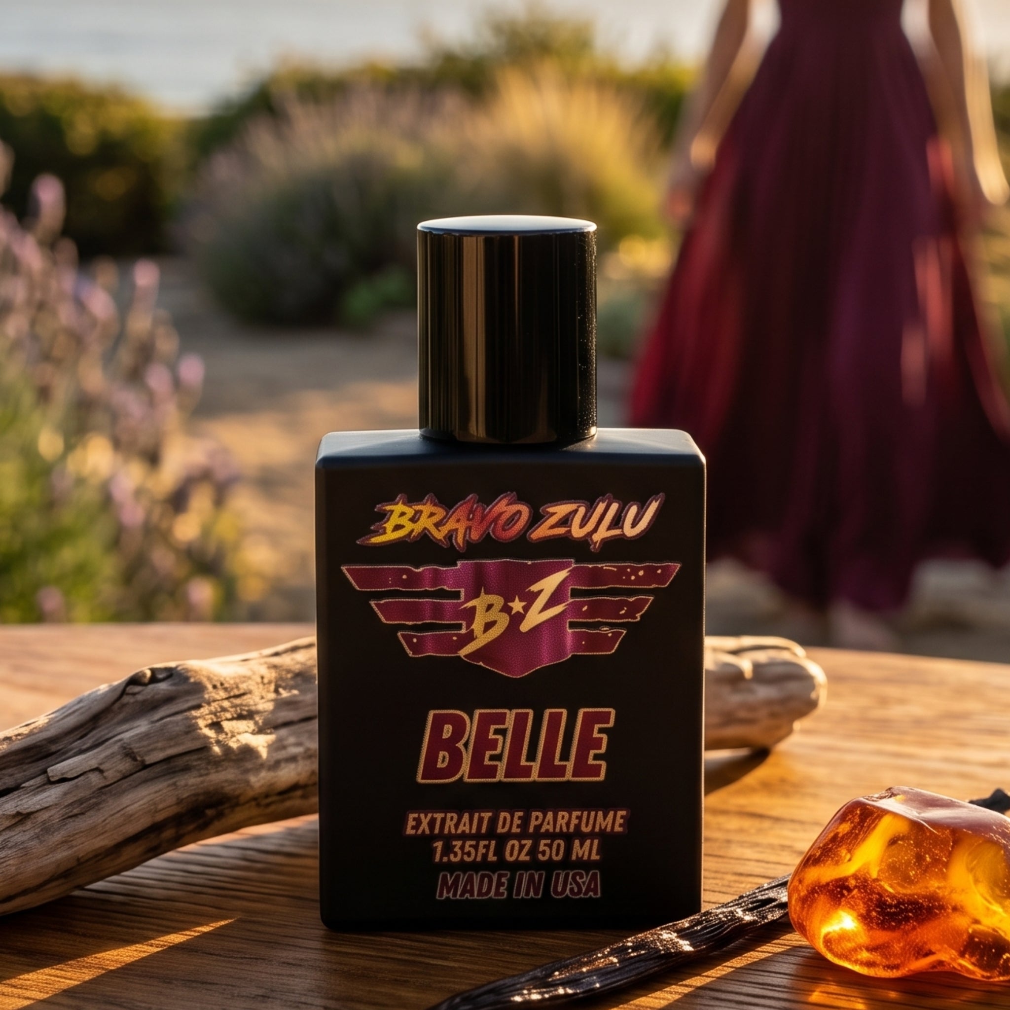 Lifestyle product shot of Bravo Zulu BELLE Extrait de Parfum by Flagship Soap Company. The matte black glass bottle with maroon-and-gold BRAVO ZULU B★Z winged logo and BELLE branding sits in sharp focus on a sun-warmed wooden table beside driftwood, a vanilla pod, and a piece of amber, with a woman in a flowing wine-colored dress standing in a sun-drenched desert garden at golden-hour sunset in the softly blurred background, evoking the fragrance's warm coconut-pineapple-vanilla character.