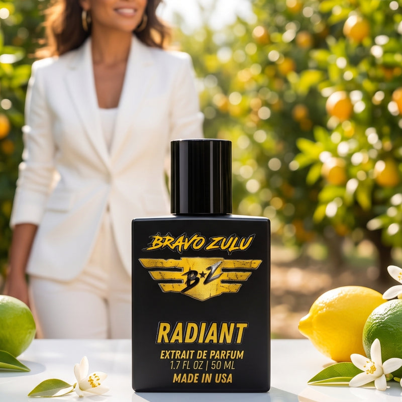 Lifestyle product shot of Bravo Zulu RADIANT Extrait de Parfum by Flagship Soap Company. The matte black glass bottle with gold-and-black BRAVO ZULU B★Z winged logo and RADIANT branding sits in sharp focus on a white table beside fresh lemons, limes, and citrus blossoms, with a poised woman in a tailored white suit standing in a sunlit citrus grove in the softly blurred background, evoking the fragrance's crisp citrus-pear-white-musk character.