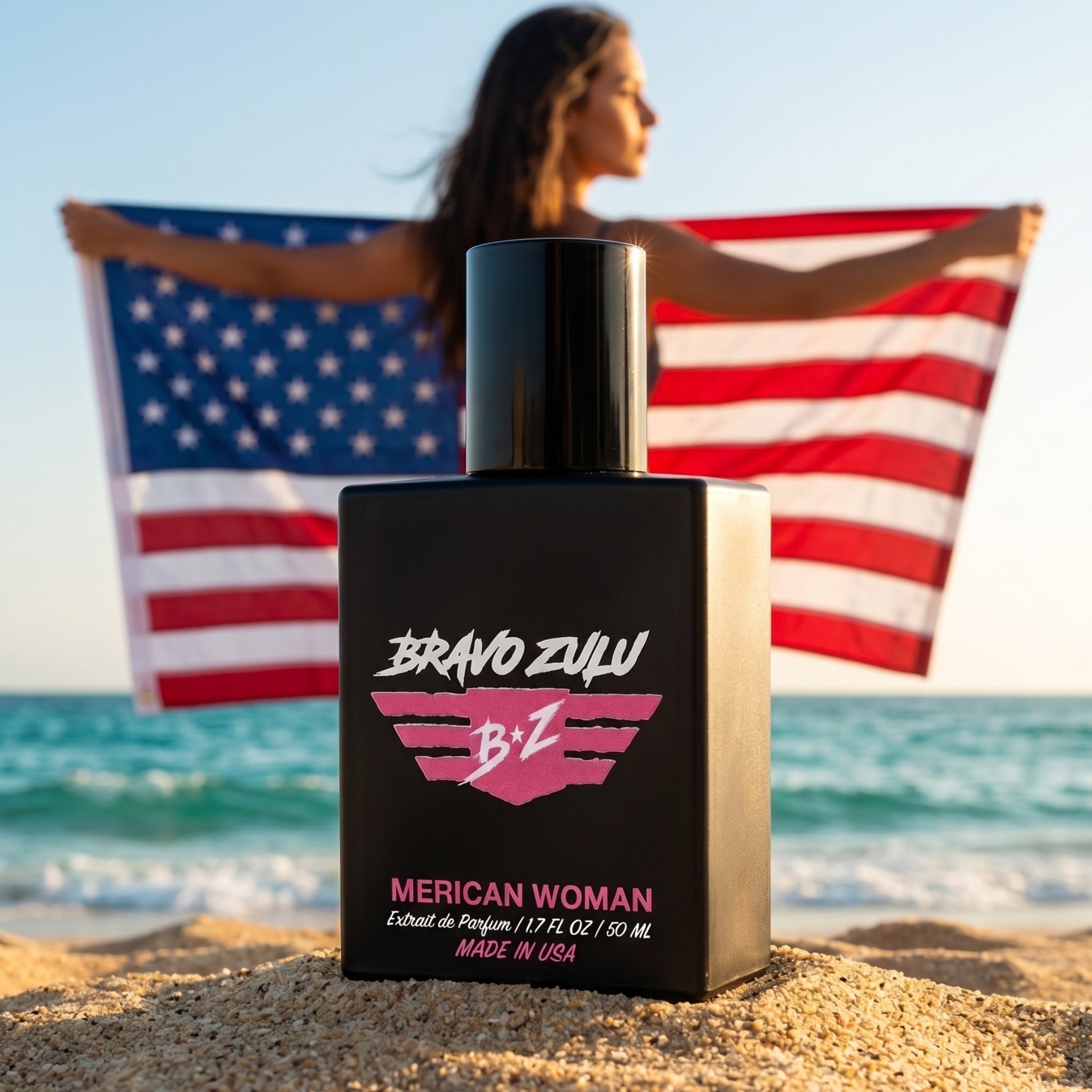 A crisp, close-up product shot of the Bravo Zulu MERICAN WOMAN Extrait de Parfum perfume bottle resting securely on a sandy beach shore. The heavy, tactical matte black glass bottle is positioned in the foreground, cleanly displaying the bold pink and white winged B★Z logo and product text. In the softly blurred background, a confident woman stands against the ocean waves at sunset, holding open a large American flag. Formulated at a high-octane 30% fragrance oil concentration and proudly made in the USA, t
