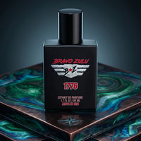 A portrait shot of Bravo Zulu's 1776 Extrait de Parfum matte black glass cologne bottle. The bottle is displayed on a tiered pedestal platform inlaid with swirling green and teal malachite stone, metallic copper trim, and an engraved compass rose. The bottle label displays the silver tactical wings logo and bold red 1776 branding, emphasizing the heritage and aggressive masculine profile of this 30% oil concentration, American-made Bay Rum fragrance.