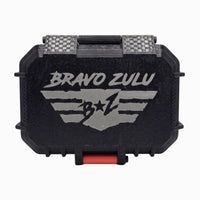 A closed Bravo Zulu tactical protective case for 1776 Extrait de Parfum, centered on a white background. The heavy-duty black case features a rugged texture, a prominent silver-gray debossed tactical wings logo, a carbon-fiber textured hinge, and a crimson red front closure latch. The premium hardware is designed to securely house and protect the 30% oil concentration, American-made masculine cologne.