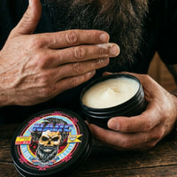 Close-up of a man with a thick, groomed beard using his finger to scoop a small amount of creamy NARC beard butter from a black tin for application.