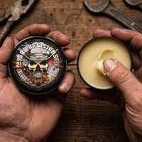 Dirty, hardworking hands opening a tin of Flagship Soap Company Bad Decisions Beard Butter. The creamy butter contains Buffalo Trace Whiskey and offers a deep whiskey and charred oak aroma for deep conditioning.