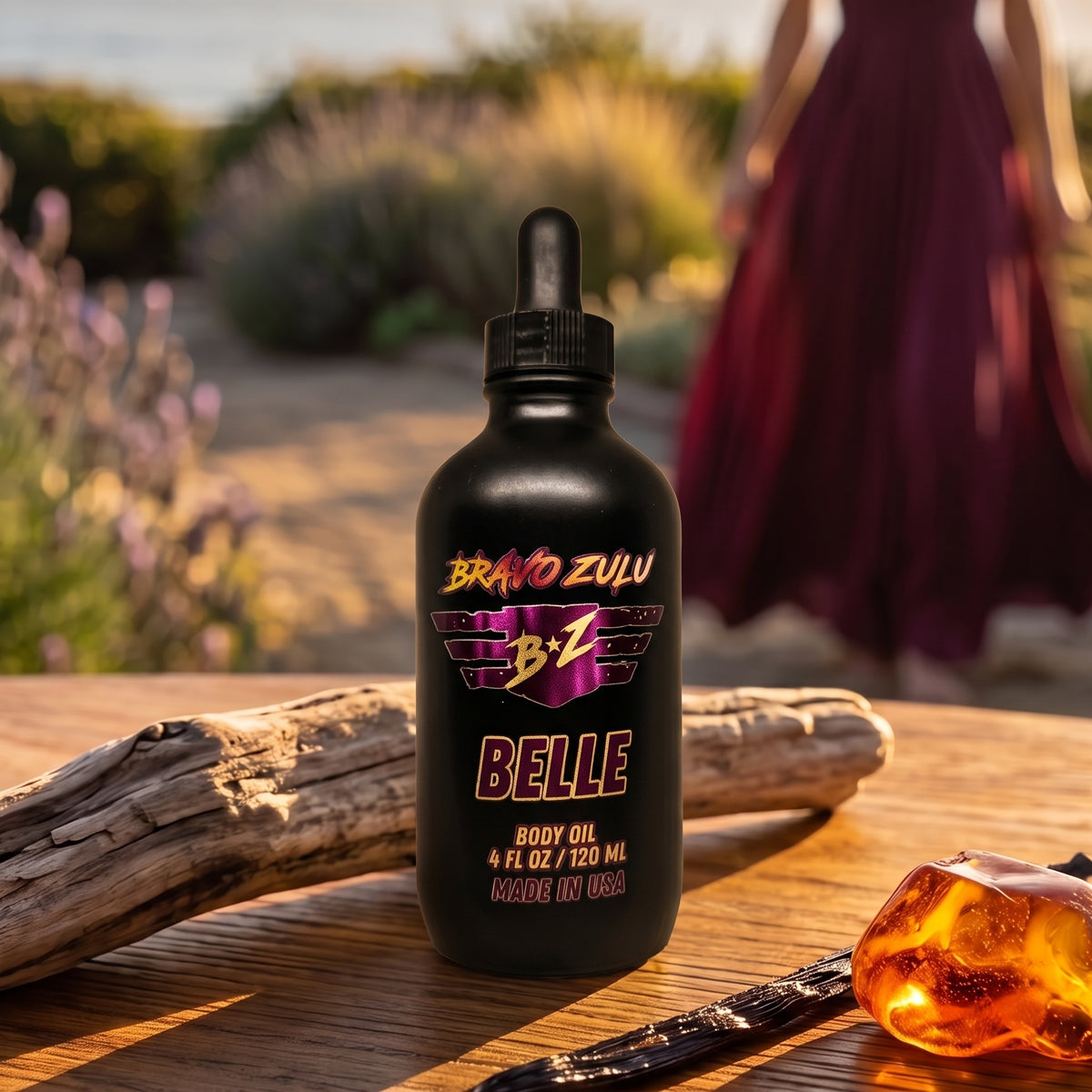 Bravo Zulu Belle Body Oil dropper bottle on a sun-warmed wooden table with driftwood, lavender, and amber stone, maroon and gold Bravo Zulu B★Z logo
