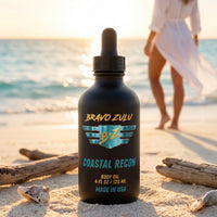 Bravo Zulu Coastal Recon Body Oil dropper bottle on a sunlit beach with driftwood and seashells, matte black glass with turquoise and gold Bravo Zulu B★Z logo