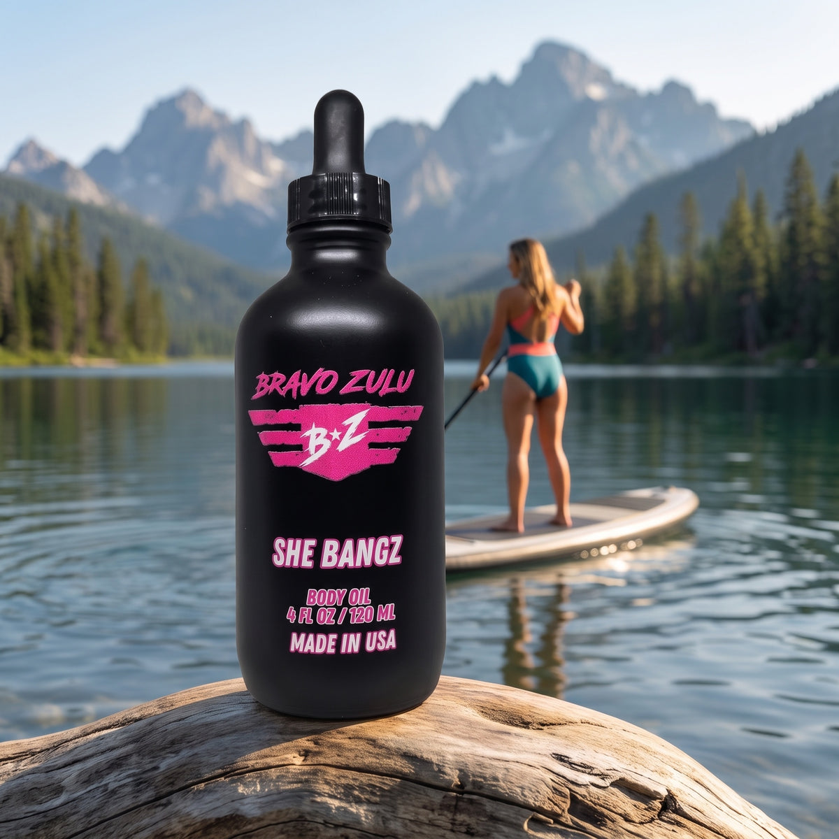 Bravo Zulu She Bangz Body Oil dropper bottle on a lakeshore with mountains and a paddleboarder in the background, pink Bravo Zulu B★Z logo