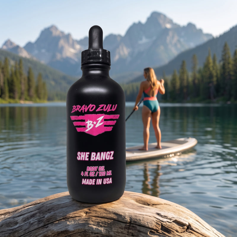 Bravo Zulu She Bangz Body Oil dropper bottle on a lakeshore with mountains and a paddleboarder in the background, pink Bravo Zulu B★Z logo