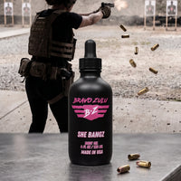Bravo Zulu She Bangz Body Oil and Extrait de Parfum bottles at an outdoor shooting range with spent brass casings, pink Bravo Zulu B★Z logo