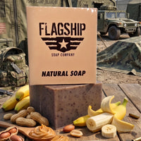 A tactical product presentation of the "Brick Maker" soap on a rough wooden military mess table. The full-package box (angled front view, showing the "SSL" acronym and crest) and the bar (back version, showing full logo and "NATURAL SOAP") are shown, surrounded by their core MRE flavor profile: a bundle of whole bananas, peanuts in the shell, a wooden spoon of peanut butter, and scattered peanuts. The background features a detailed military camp with olive-drab canvas tents 