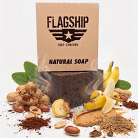 Flagship Soap Company "Brick Maker" box (the back version, showing the full "FLAGSHIP SOAP COMPANY" and "NATURAL SOAP" text) is positioned with its fresh ingredient profile: ripe bananas (whole, sliced, and peeled), whole peanuts in the shell, peanut butter spread, cocoa powder, and walnut shells (whole and crushed). Highlighting the moisturizing formulation (Coconut, Palm, Avocado oils, Shea Butter) and its medium exfoliation grit (walnut shells, cocoa). A deep-cleansing and hydrating bar