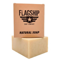 A studio product shot of Holy Grail Natural Soap by Flagship Soap Company. The tan packaging features a bold, stenciled military-style logo with a star and wings, reminiscent of a Meals Ready to Eat (MRE) ration. The box sits atop a thick, artisan-cut bar of cream-colored natural soap against a white background. Designed with a rugged aesthetic, this MRE themed soap makes an excellent gift for any veteran, active-duty service member, or first responder who values high-quality, American-made skin care.