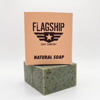 Vertical product shot of 'Flagship Soap Company' natural exfoliating bar (green with coffee specks) stacked beneath its peach-colored cardboard box on a white background. The box displays the 'Flagship' logo, 'NATURAL SOAP', and 'MRE-THEMED GIFT SOAP' text. The underlying soap bar's texture is clearly visible, showcasing the coffee and sea clay exfoliating particles. High-performance, patriotic veteran gift soap from Flagship Soap Company.