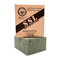 Vertical product shot featuring the 'Flagship' 'Shitlets-Spearmint' exfoliating soap bar in its packaging box stacked on top of an unwrapped bar. The peach-colored box clearly displays the 'SSL' (Shit-Shower-Lather) brand, 'Natural Soap Bar', 'Spearmint' scent, 'Warfighter Approved' endorsement, and '5.56 OZ' weight. The unwrapped green bar shows its granular texture with dark coffee specks. High-performance, patriotic veteran gift soap from Flagship Soap Company, made in Idaho, USA.