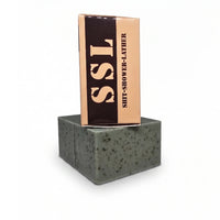 Vertical, front view of 'Flagship' 'Shitlets' (MRE spearmint and coffee natural soap bar) and its cardboard packaging box on a white background. The box displays the large vertical text 'SSL' and 'SHIT-SHOWER-LATHER'. The underlying soap bar is green with visible exfoliating coffee specks and sea clay texture. High-performance, patriotic veteran gift soap from Flagship Soap Company.