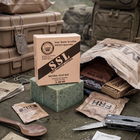 Front view of 'Flagship' Shitlets-Spearmint MRE-themed exfoliating soap bar and its cardboard packaging box placed on a tactical green field jacket. The bar is green with visible coffee specks. The package displays the 'Flagship' seal, the text 'SHIT-SHOWER-LATHER', 'NATURAL SOAP BAR', and '5.56 OZ'. The soap is surrounded by actual MRE components: a bag with 'BEEF STEW' visible, an 'FRH HEATER POUCH', a pack of 'MRE COFFEE MIX', and a mult-itool. The background features military gear bags and hard cases wi